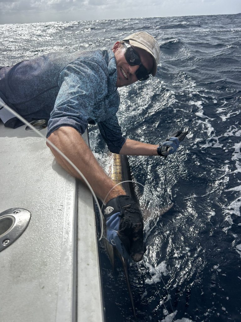 Lachend bemanningslid leunt over de reling, gehandschoende hand aan de snuit van een billfish, en zet hem terug tijdens een visreis op Bonaire
