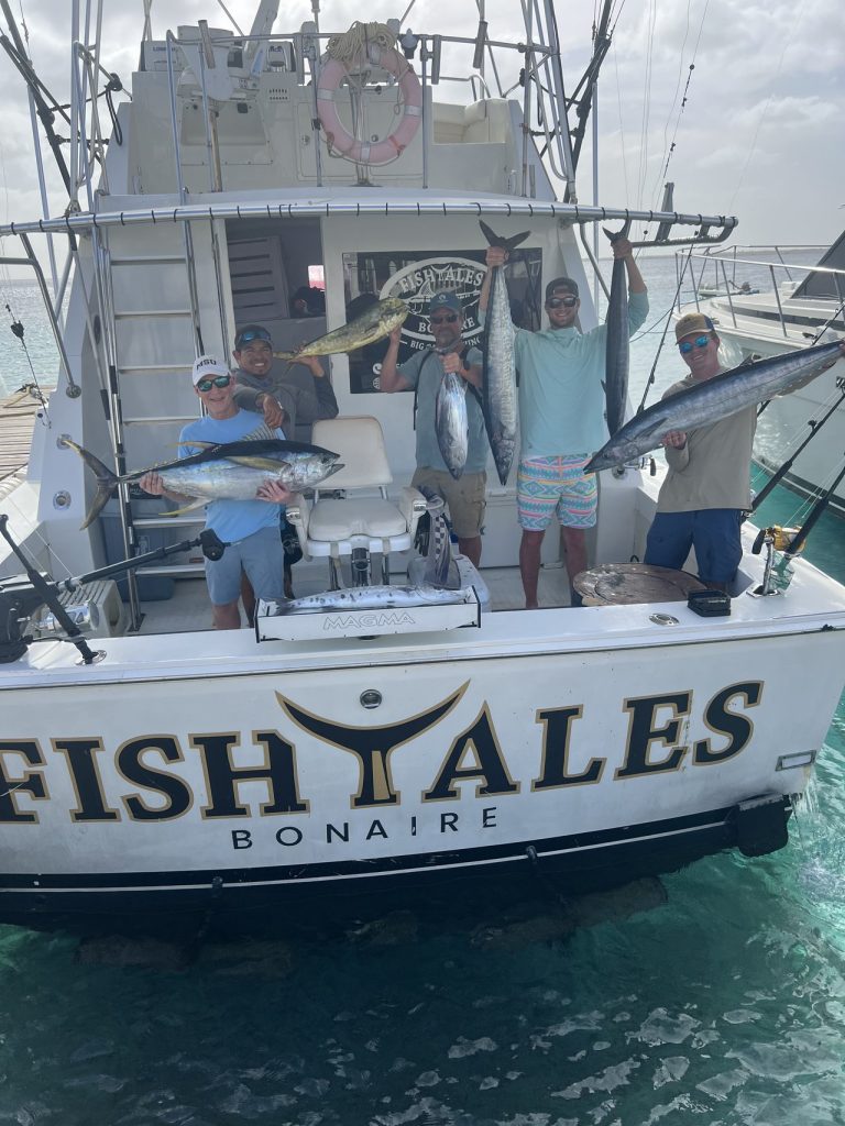 Vijf sportvissers op de Fishtales Bonaire boot houden een gemengde vangst van tonijn, wahoo en mahi-mahi omhoog aan het achterdek.