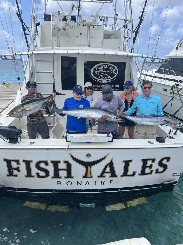 Zes gasten houden tonijn en lange wahoo omhoog aan het achterdek van de Fishtales Bonaire sportvisser, met de naam FISH TALES op de spiegel.