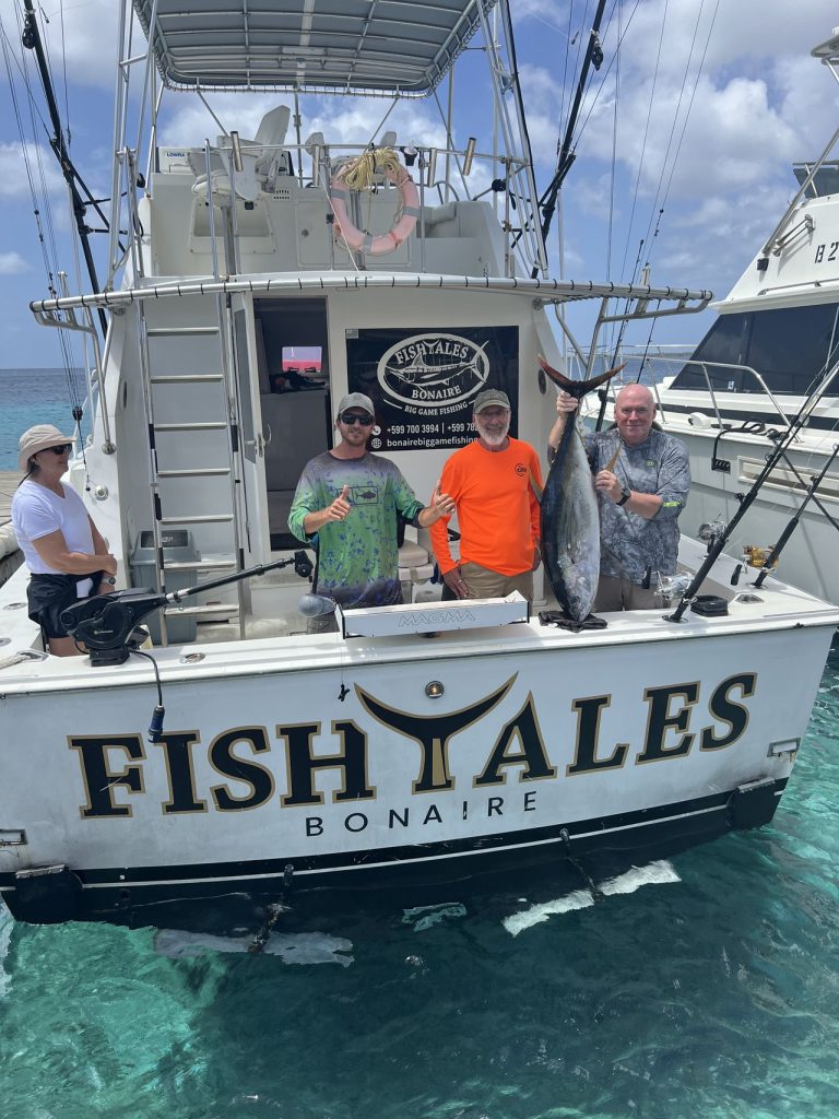 Sportvissers en bemanning aan het achterdek van de Fishtales Bonaire boot, een houdt een grote donkere tonijn vast naast een naburige sportvisser.