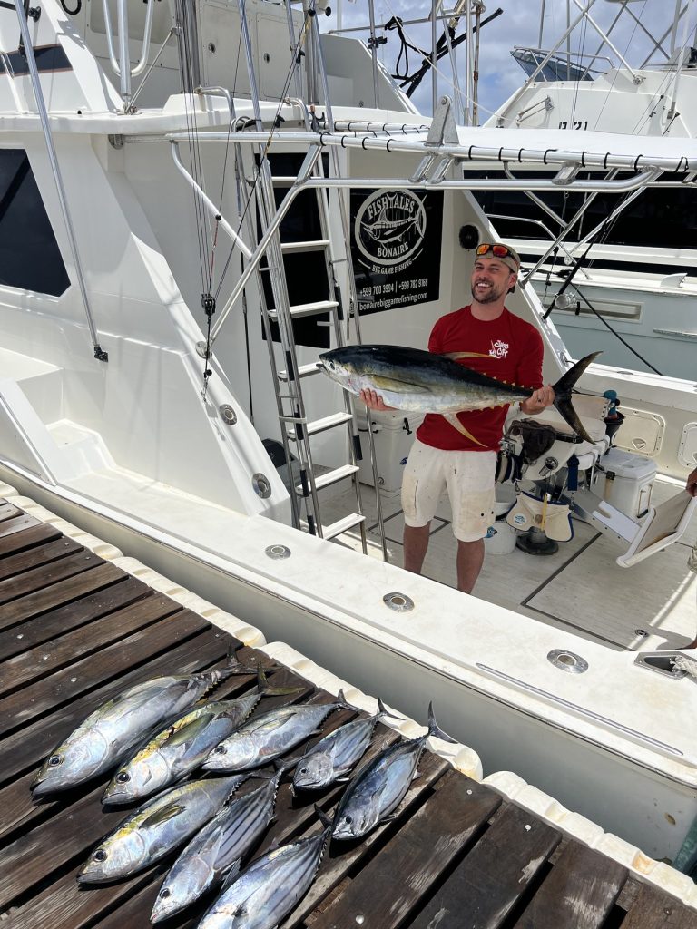 Een rij tonijn ligt op de kade terwijl een sportvisser aan boord van de Fishtales Bonaire boot op de achtergrond nog een tonijn omhoog houdt.