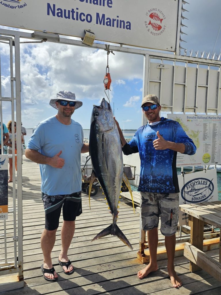 Twee vissers steken hun duimen op terwijl ze een tonijn vasthouden bij de steiger van Bonaire Nautico Marina na een Fishtales-visserijcharter.