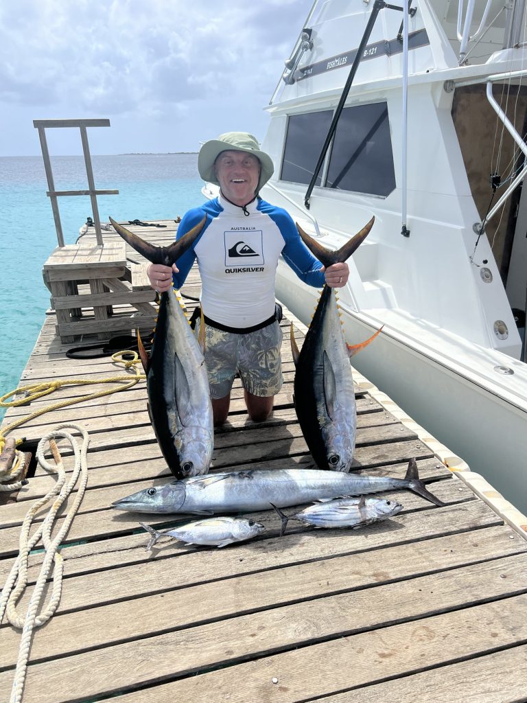 Visser met een zonnehoed houdt twee grote tonijnen vast met een kleinere tonijn op de steiger naast de Fish Tales Bonaire boot