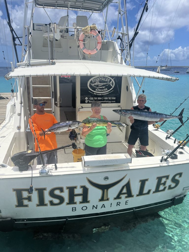 Drie vissers op de Fish Tales Bonaire boot houden een tonijn, een gouden mahi-mahi en een wahoo vast over de spiegel in blauw water.