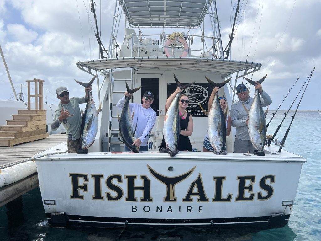 Groep sportvissers aan het achterdek van de Fishtales Bonaire boot houdt meerdere geelvintonijnen omhoog boven de gouden FISH TALES spiegel.