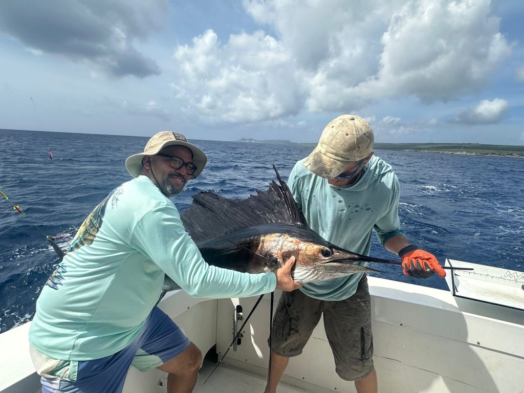 Twee lachende bemanningsleden houden een zeilvis vast langs de boot aan dek met de kustlijn van Bonaire en blauwe lucht achter hen.