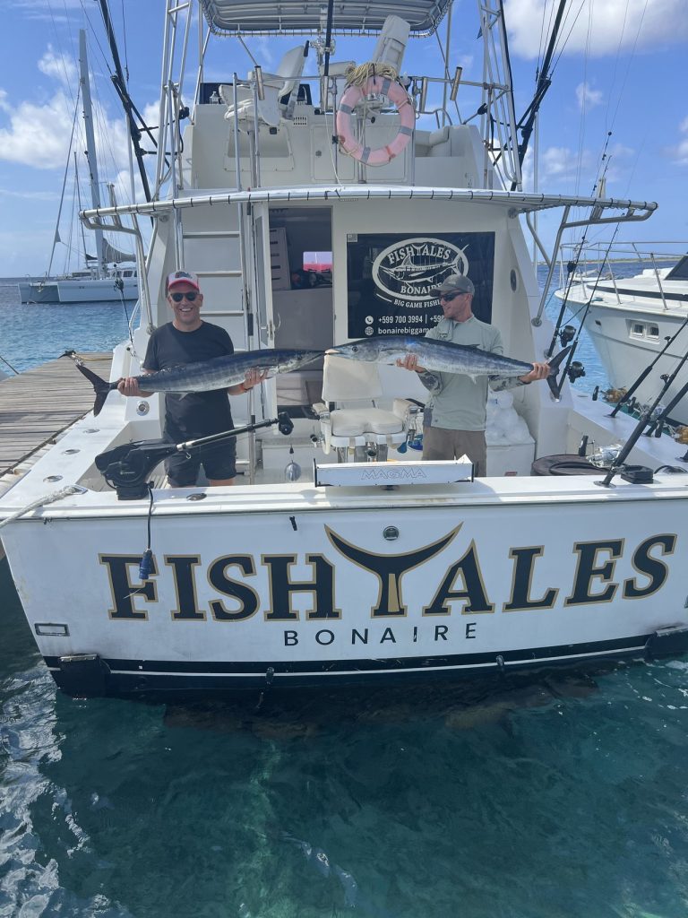 Twee vissers staan op de Fishtales Bonaire-sportvisser met grote wahoo boven de spiegel met opschrift FISH TALES BONAIRE bij de jachthaven.
