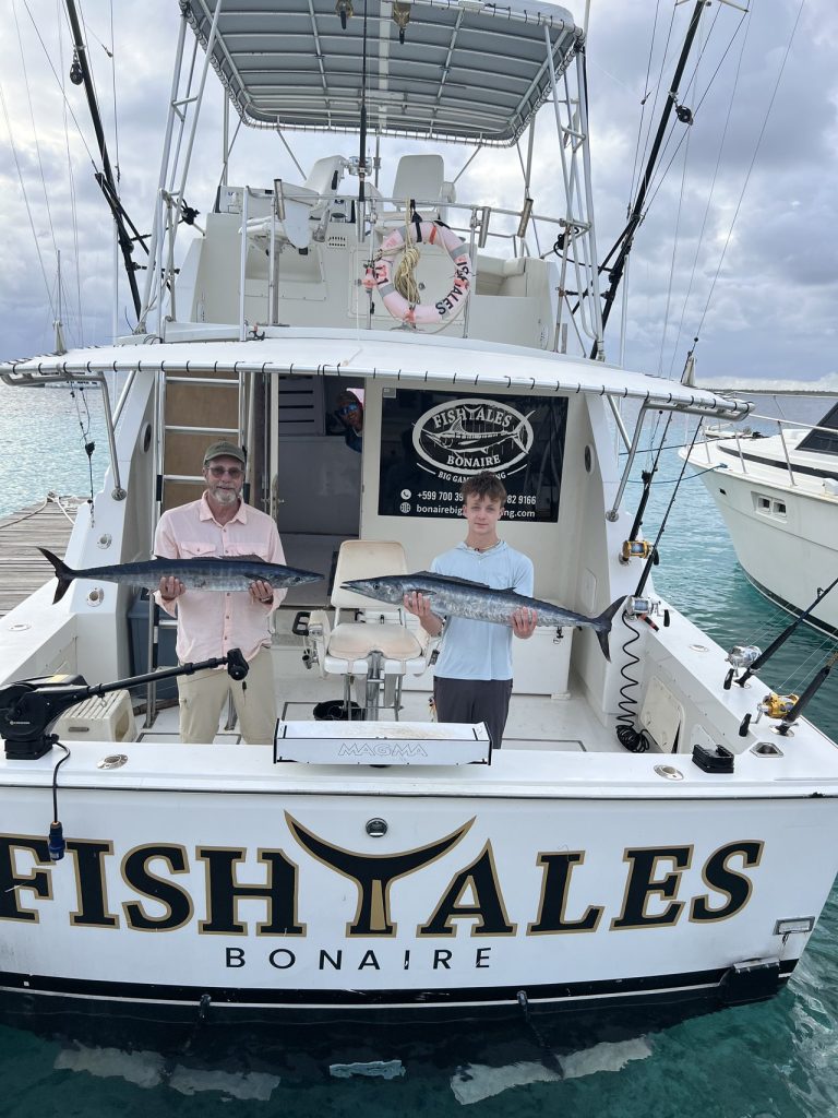 Twee vissers staan in de kuip van de Fish Tales Bonaire-sportvisser, elk met een wahoo, achter de gouden spiegel bij de steiger.