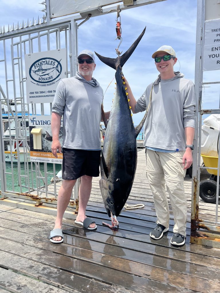 Twee sportvissers staan naast een hangende geelvintonijn op de houten kade bij de Fishtales Bonaire banner bij Nautico Marina, Kralendijk.