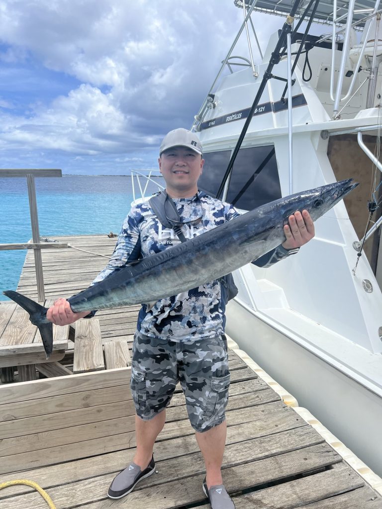 Lachende visser in camoshirt houdt een lange wahoo vast op de houten steiger naast de Fish Tales sportvisser in blauw water.
