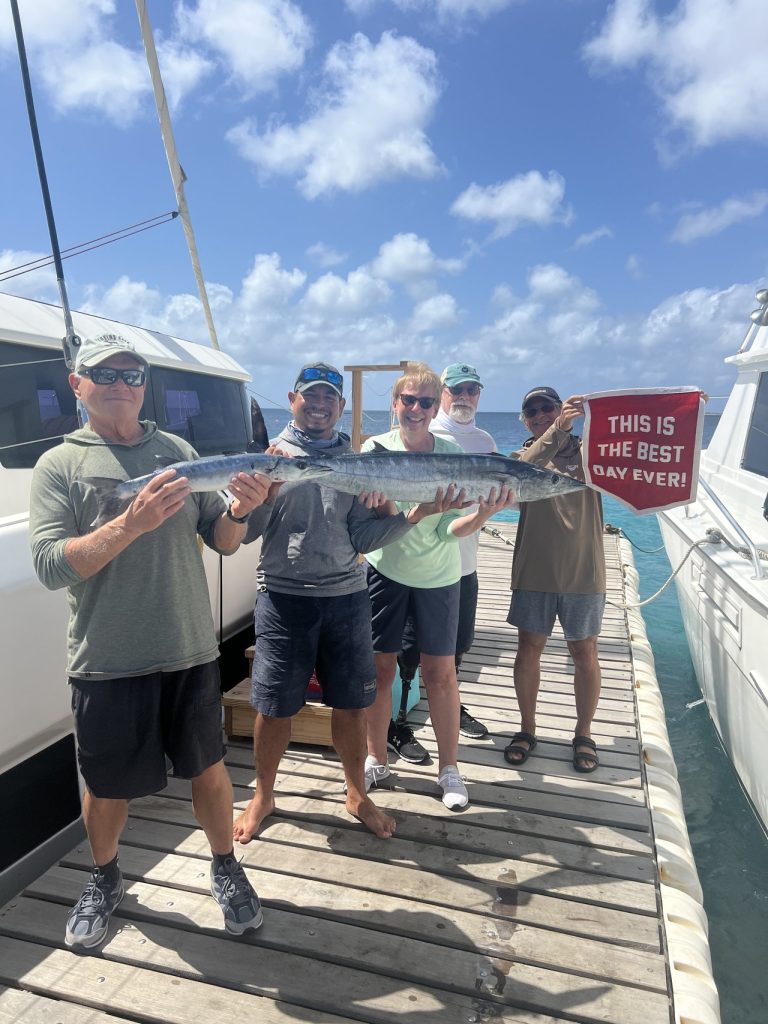 Twee sportvissers houden een lange wahoo vast op de kade terwijl een groep achter hen zwaait met een rode vlag met 'This Is The Best Day Ever' op Bonaire.
