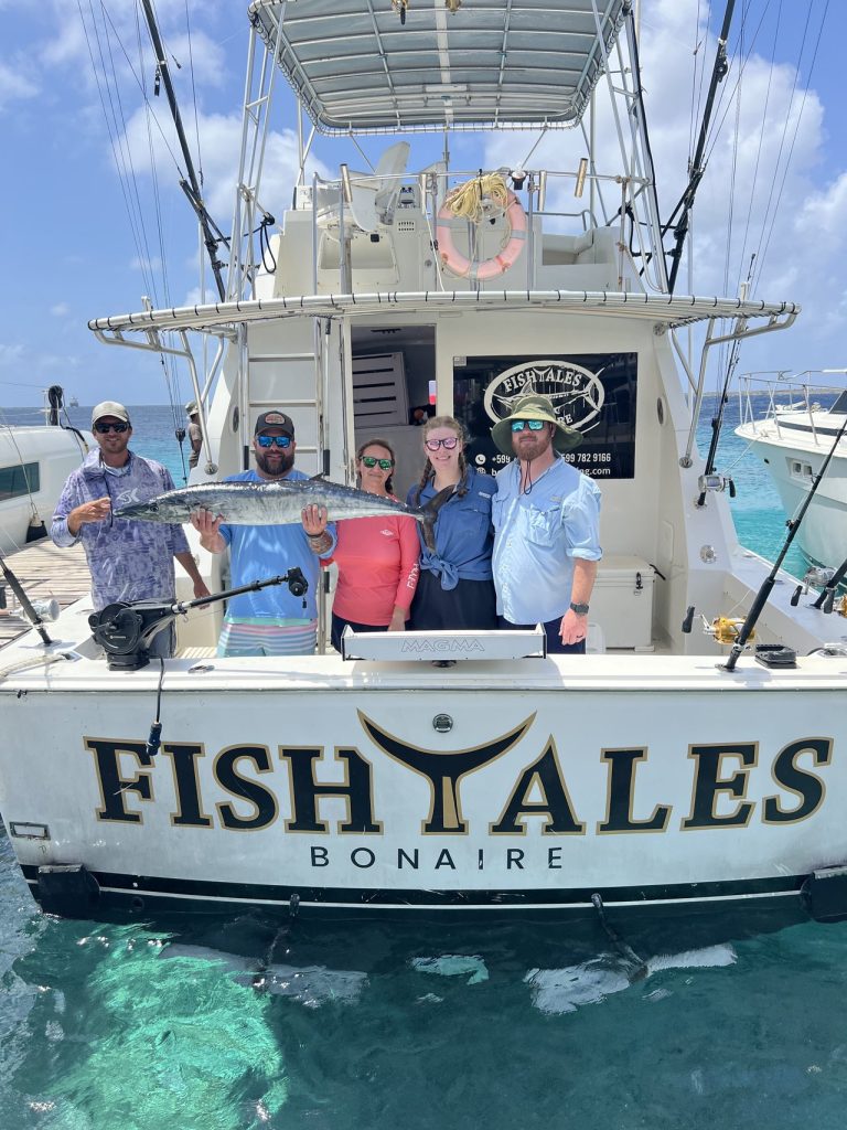 Vijf sportvissers aan het achterdek van de Fishtales Bonaire boot, een houdt een zilveren wahoo omhoog boven de FISH TALES spiegel boven helder water.