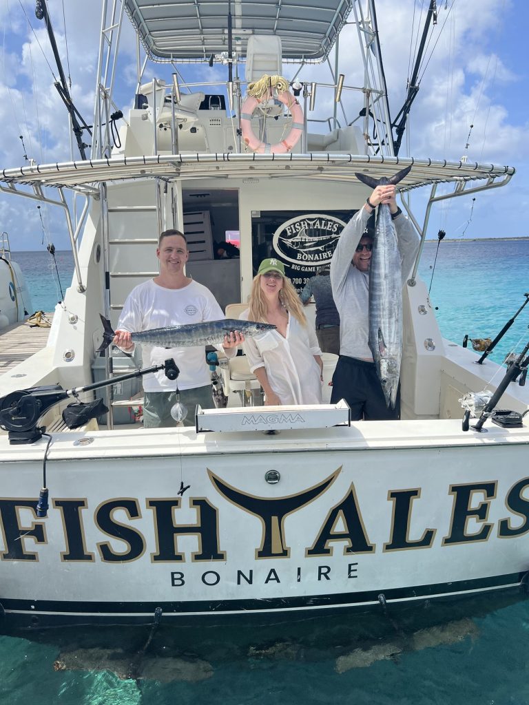 Drie sportvissers op de Fishtales Bonaire sportvisser houden twee lange wahoo vast aan het achterdek boven turquoise Caribisch water.