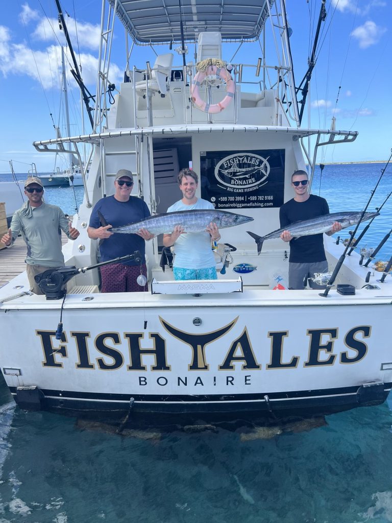 Vier vissers aan boord van de Fishtales Bonaire-sportvisser met twee grote wahoo boven de spiegel met opschrift FISH TALES BONAIRE.