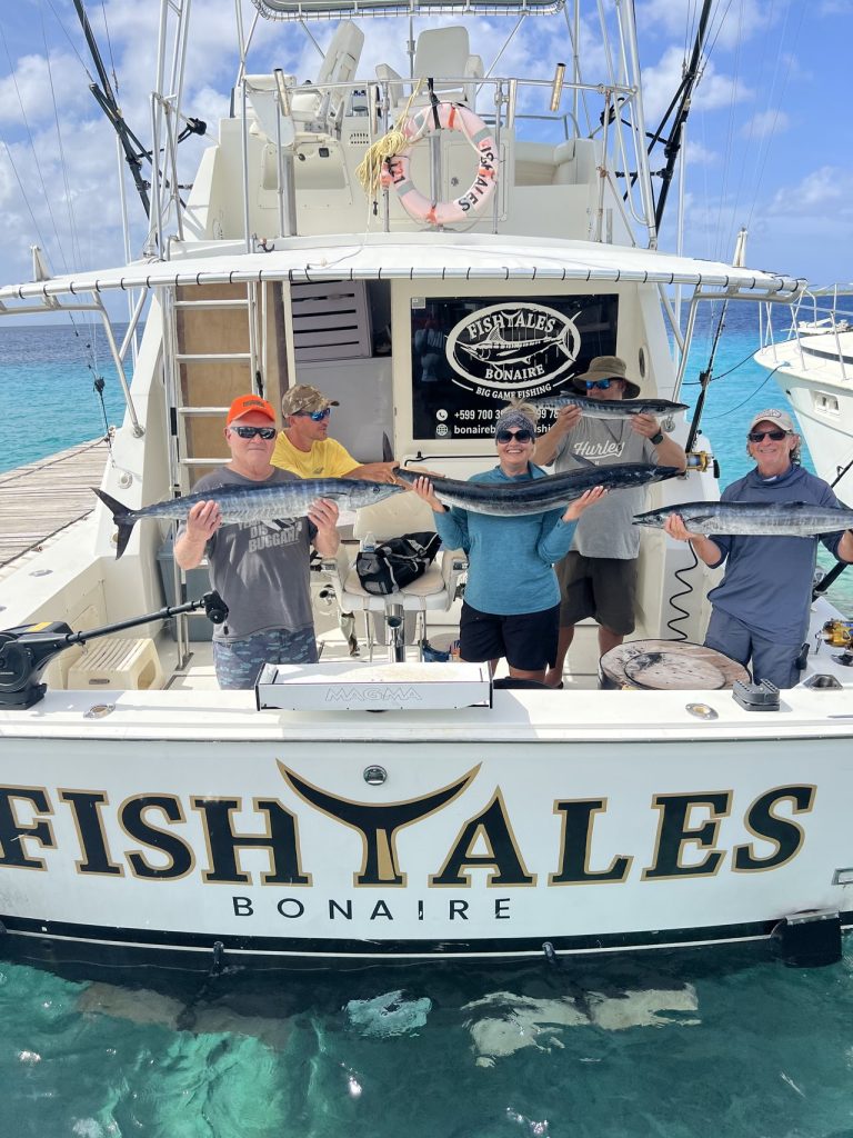 Vijf vissers houden lange wahoo vast op de achtersteven van de Fishtales Bonaire-boot boven helder turquoise water met de marina op de achtergrond.