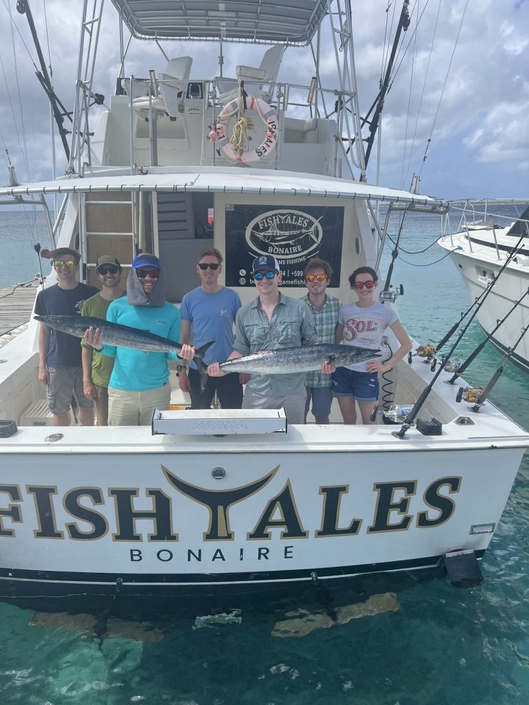 Groep van zeven vissers houdt twee lange wahoo vast op de achtersteven van de Fish Tales Bonaire-sportvissersboot in turquoise water.