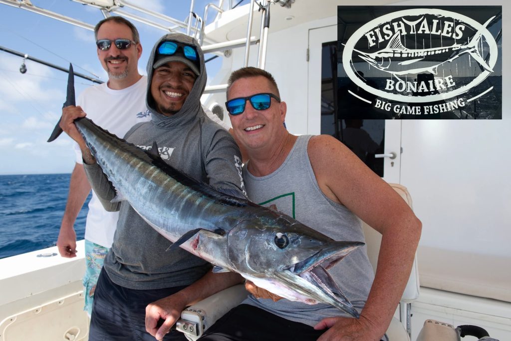 Drie lachende mannen houden een grote wahoo vast aan boord van de Fishtales boot, met het Fishtales Bonaire Big Game Fishing logo in de hoek.