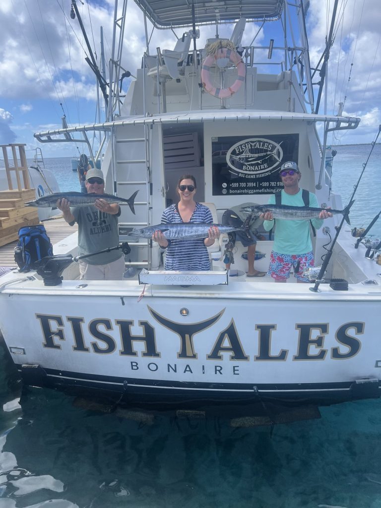 Drie gasten aan boord van de Fishtales Bonaire-sportvisser, elk met een wahoo boven de spiegel met opschrift FISH TALES BONAIRE.