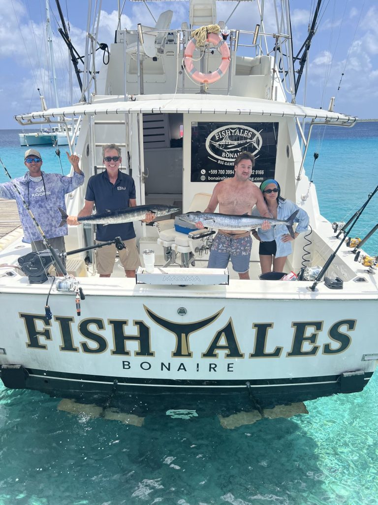Vissers en bemanning op de spiegel van de Fish Tales Bonaire houden twee grote wahoo vast boven het heldere turquoise Caribische water.
