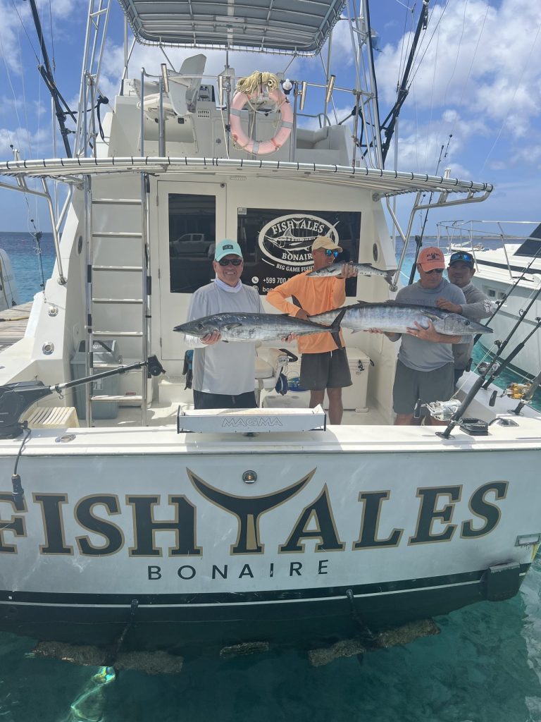 Vier sportvissers staan aan het achterdek van de Fishtales Bonaire boot en houden meerdere grote wahoo omhoog boven de FISH TALES spiegel.