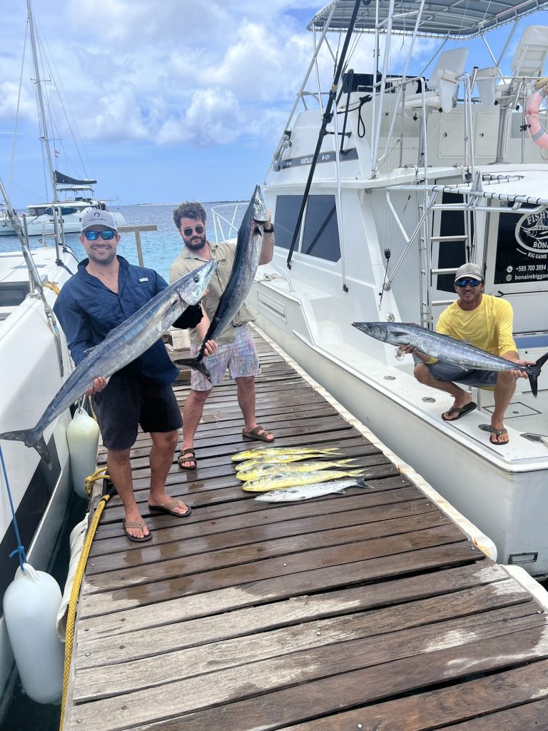 Anglers on the dock at Bonaire's marina holding wahoo and tuna beside the Fishtales boat, with mahi-mahi laid out on the planks.
