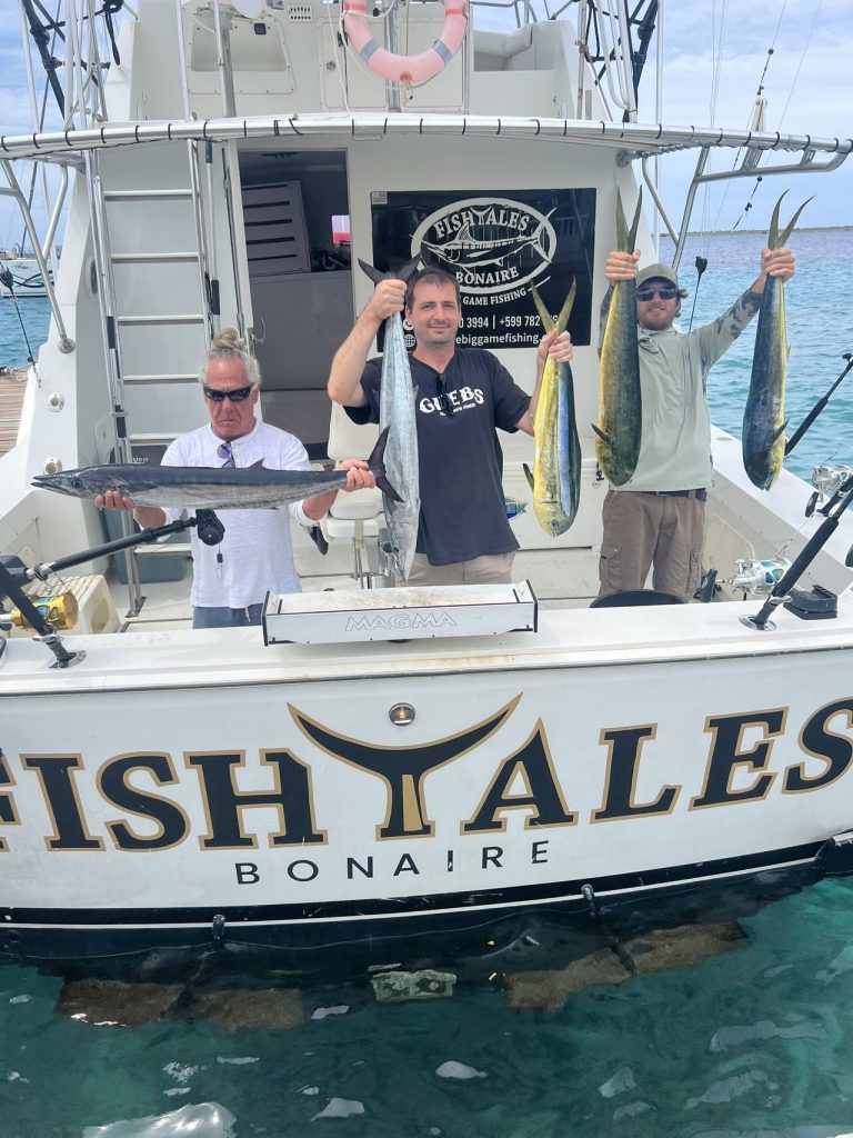 Drie vissers houden een wahoo en meerdere mahi-mahi omhoog op de spiegel van de Fish Tales Bonaire-boot boven helder turquoise water.