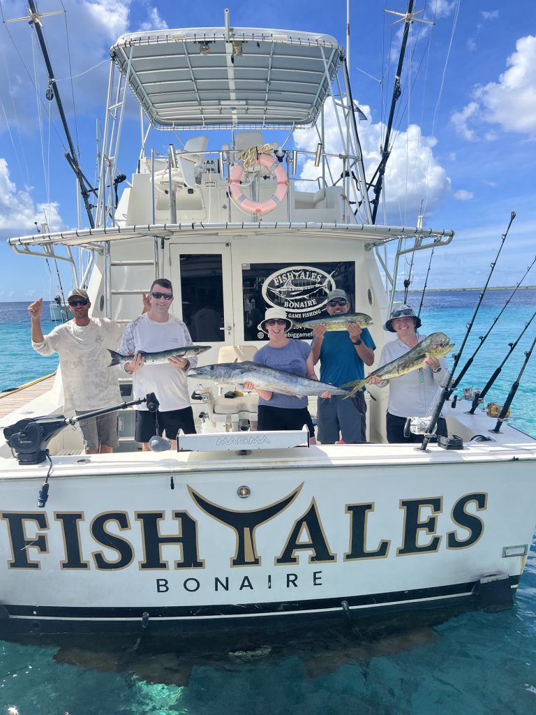 Vijf vissers en bemanning op de Fishtales Bonaire-sportvisser met wahoo en mahi-mahi boven turquoise water, FISH TALES-spiegel.
