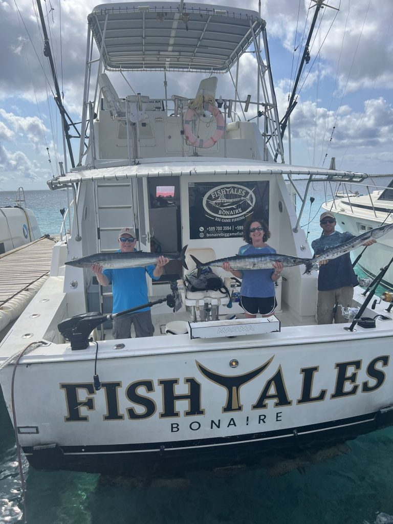 Drie sportvissers houden wahoo vast op het achterdek van de Fishtales Bonaire charterboot, afgemeerd in helder turquoise water.
