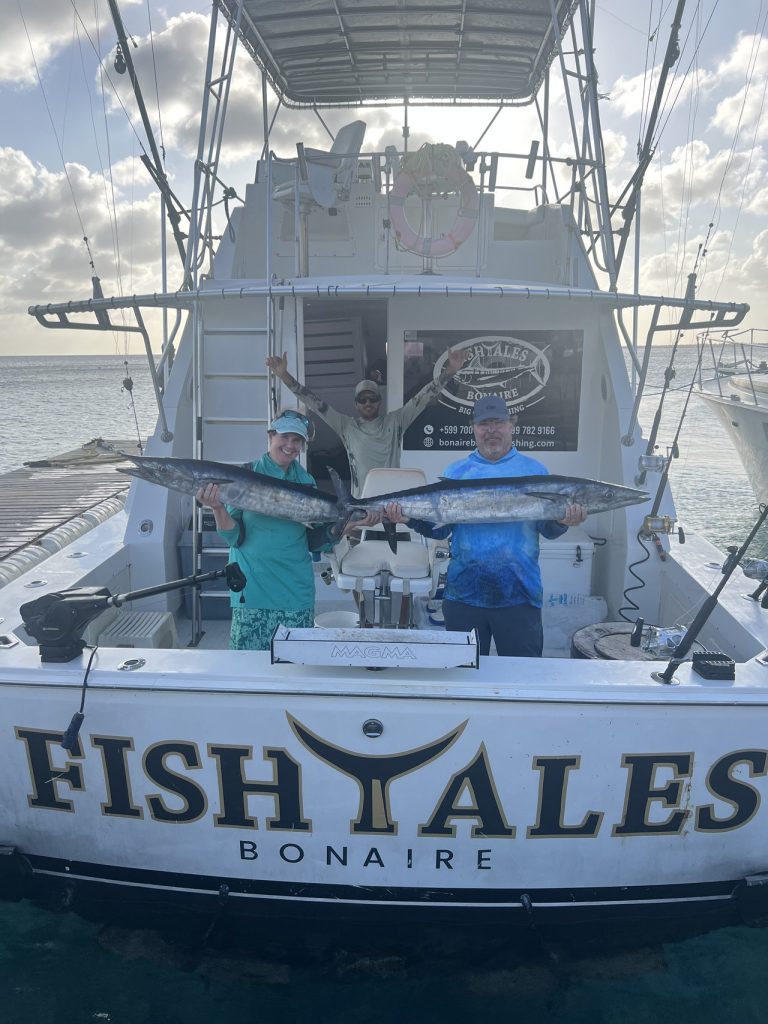 Twee sportvissers houden een paar lange wahoo vast aan het achterdek van de Fishtales Bonaire boot onder een zachte avondlucht.