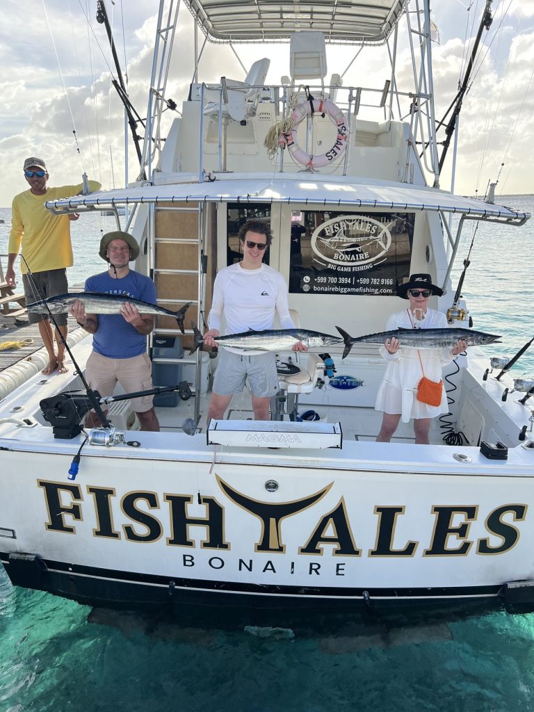 Vier vissers houden drie wahoo vast over de achtersteven van de Fish Tales Bonaire charterboot aan de steiger in helder Caribisch water