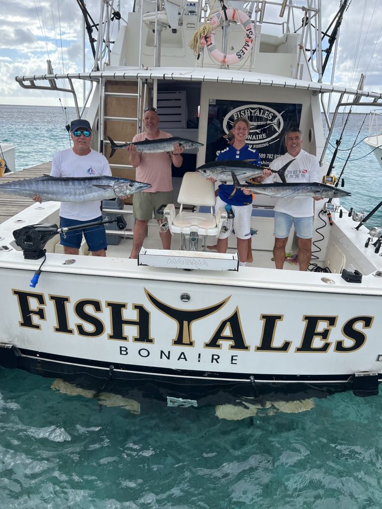 Vier vissers houden wahoo en tonijn omhoog op de achtersteven van de Fishtales Bonaire-sportvissersboot boven turquoise rifwater.