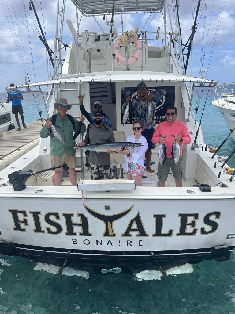 Vijf sportvissers aan het achterdek van de Fishtales Bonaire boot houden een wahoo en meerdere tonijnen vast boven de FISH TALES spiegel en helder water.