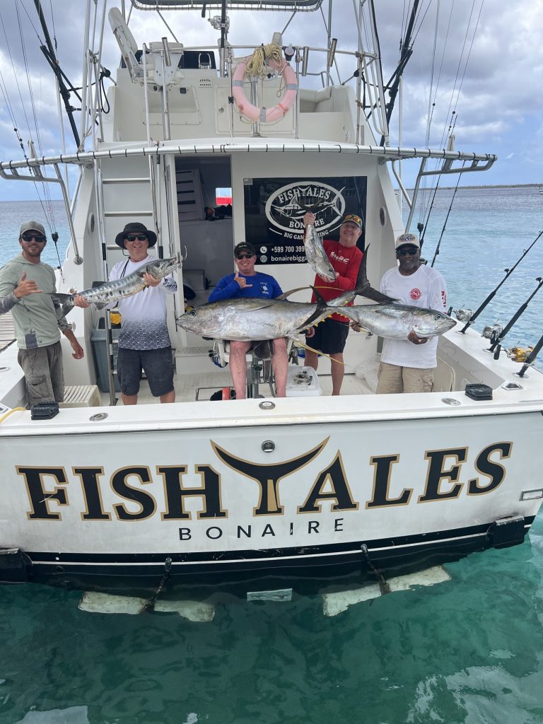 Vijf sportvissers aan het achterdek van de Fishtales Bonaire boot houden wahoo en grote geelvintonijn omhoog boven de gouden FISH TALES spiegel.