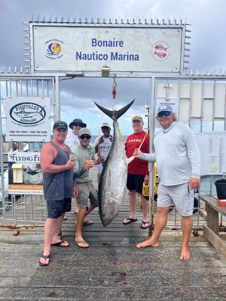 Vijf sportvissers poseren met een grote geelvintonijn die onder het bord van Bonaire Nautico Marina hangt aan de Fishtales-kade in Kralendijk.