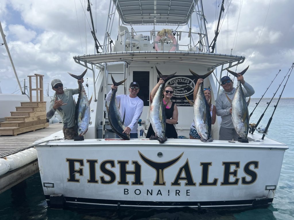Vijf vissers aan de achtersteven van de Fishtales Bonaire-boot houden een rij geelvintonijnen omhoog boven het rustige marinawater
