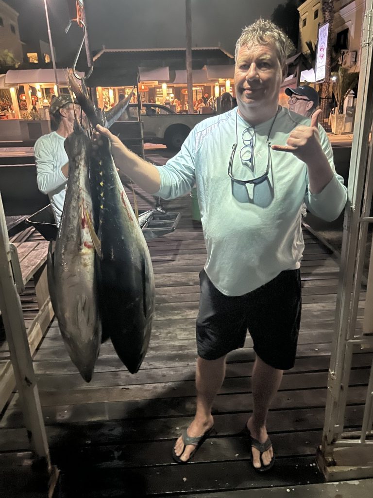 Lachende visser die een shaka maakt terwijl hij twee grote tonijnen vasthoudt die 's nachts aan het fileerrek in de jachthaven van Bonaire hangen