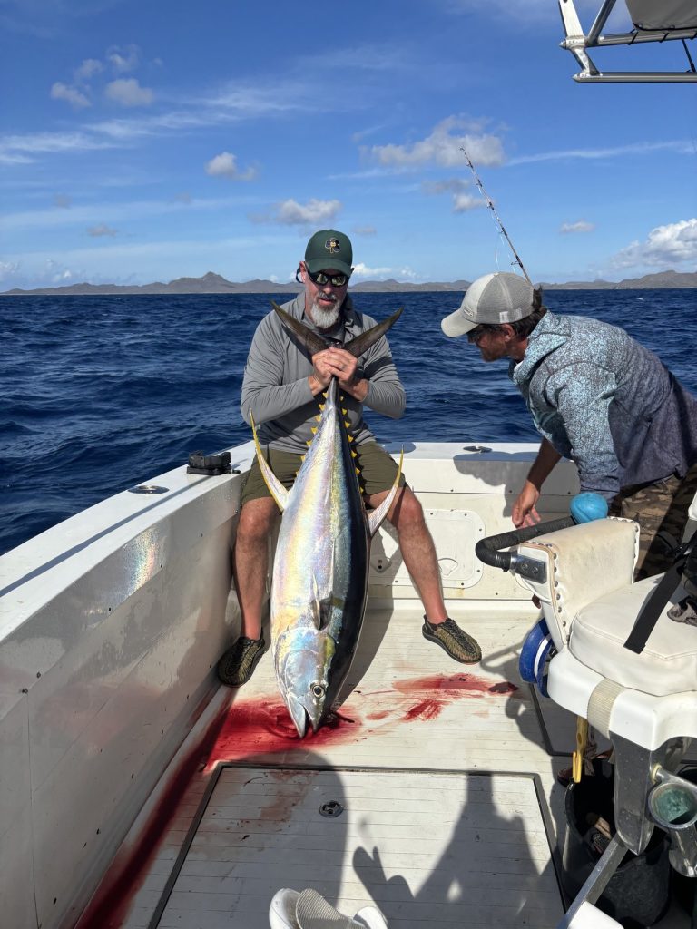 Visser houdt een grote geelvintonijn aan boord vast terwijl een bemanningslid van Fishtales helpt, kobaltblauwe Caribische zee op de achtergrond.