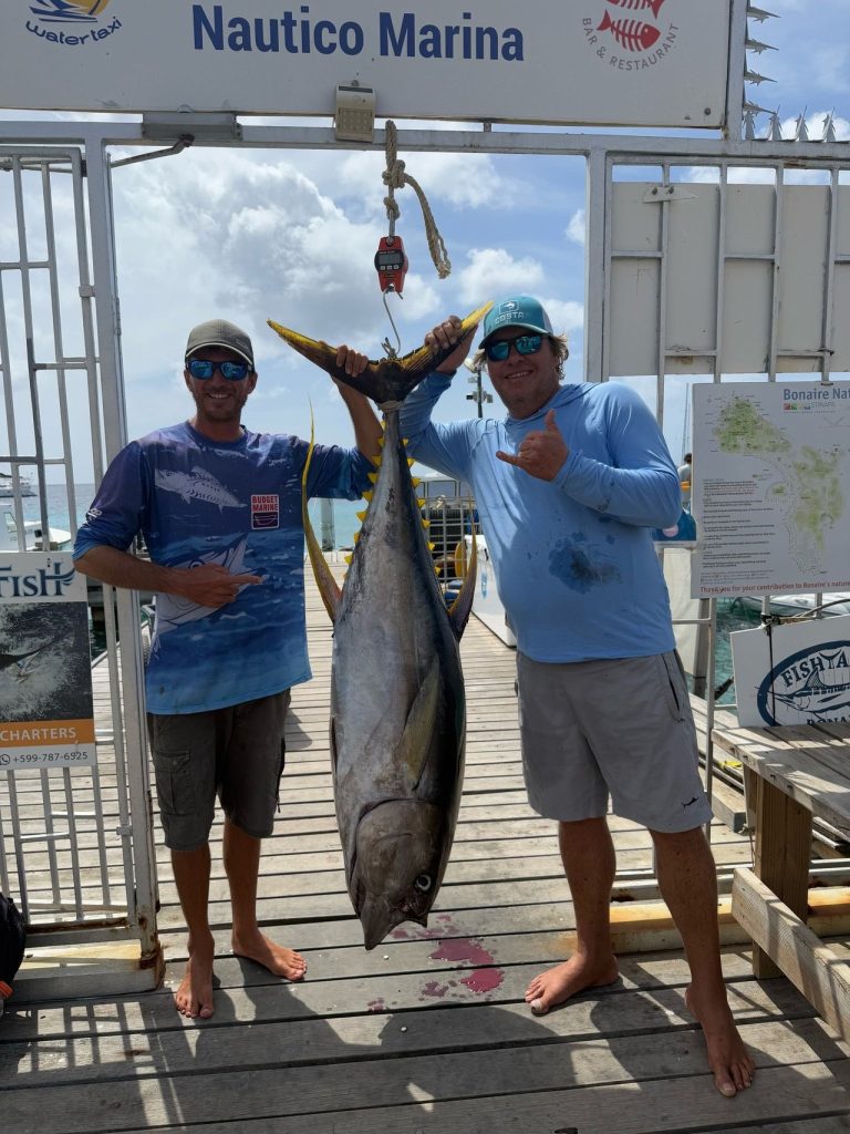 Twee vissers poseren met een grote geelvintonijn aan een weegschaal onder het bord van Nautico Marina in Kralendijk, Bonaire.