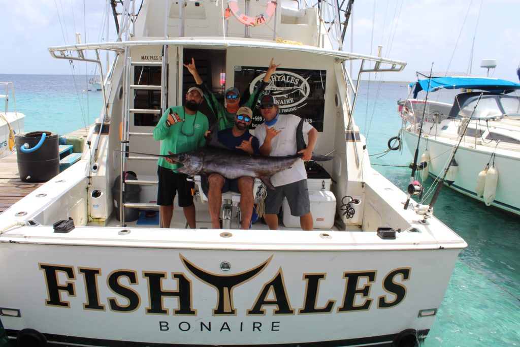 Fishtales Bonaire-bemanning met een zwaardvis op de achtersteven van de boot, turquoise water op de achtergrond
